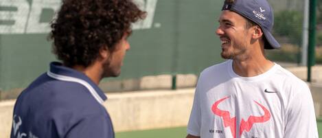 Dos personas en una cancha de tenis, con un muro verde al fondo.