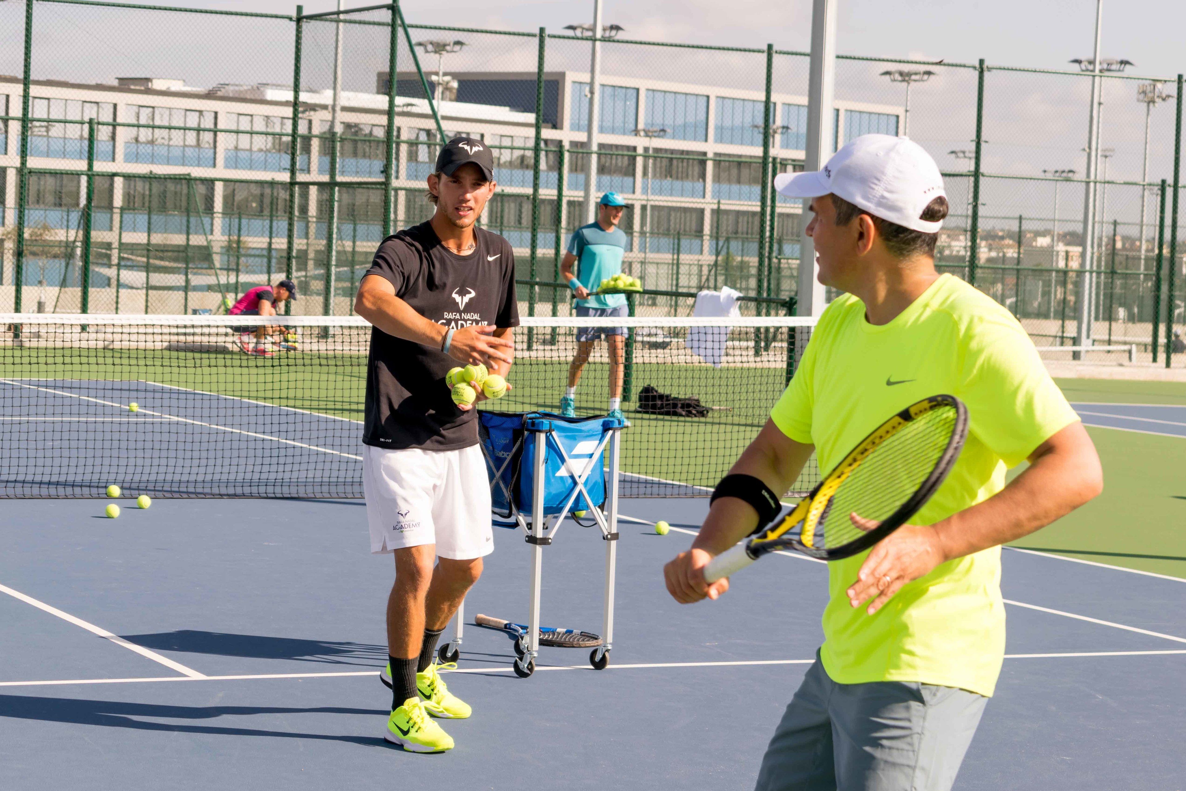 Una cancha de tenis con jugadores, una red y un carrito para pelotas.