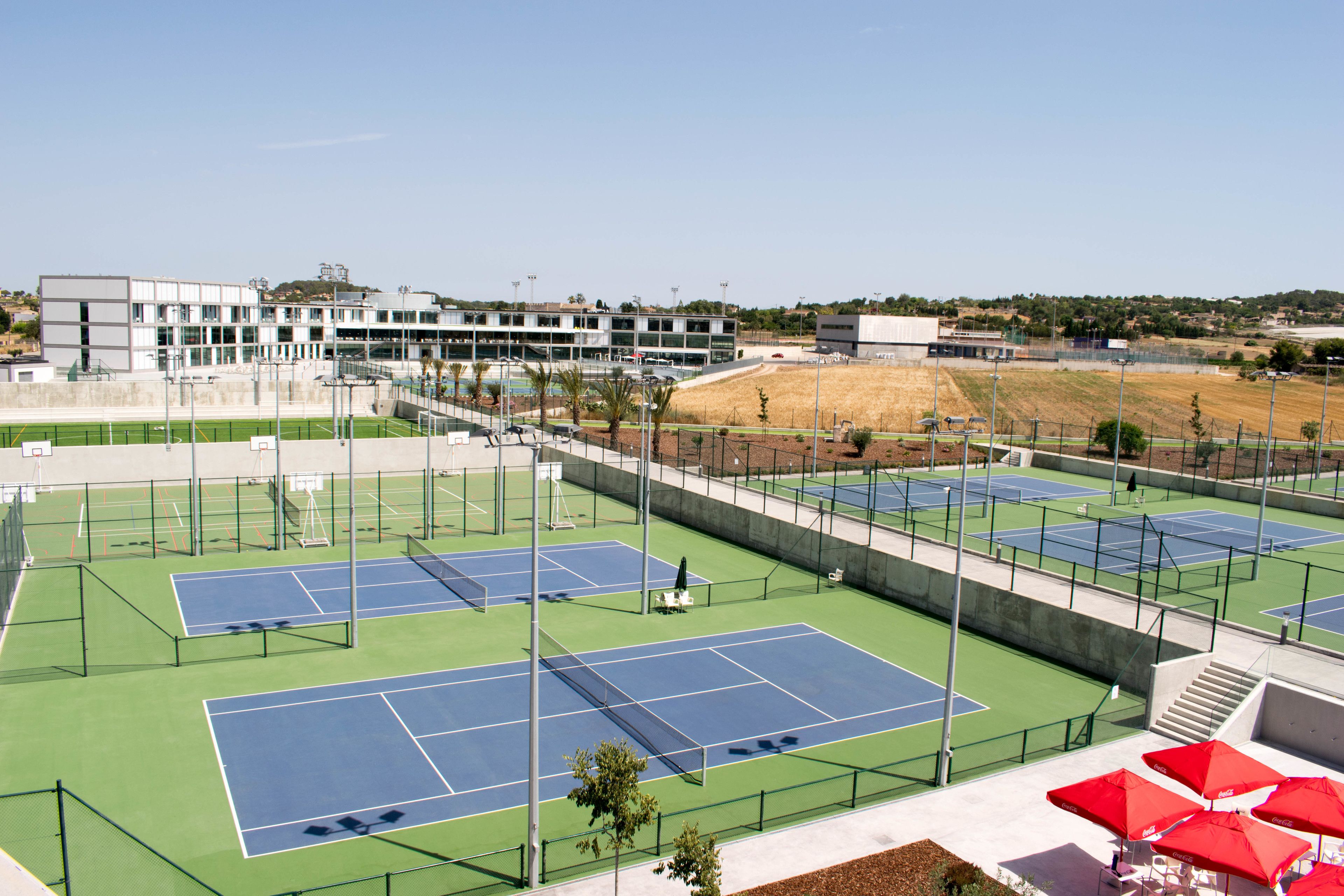 Un complexe de courts de tennis comprenant plusieurs terrains, un bâtiment en arrière-plan et des parasols rouges.