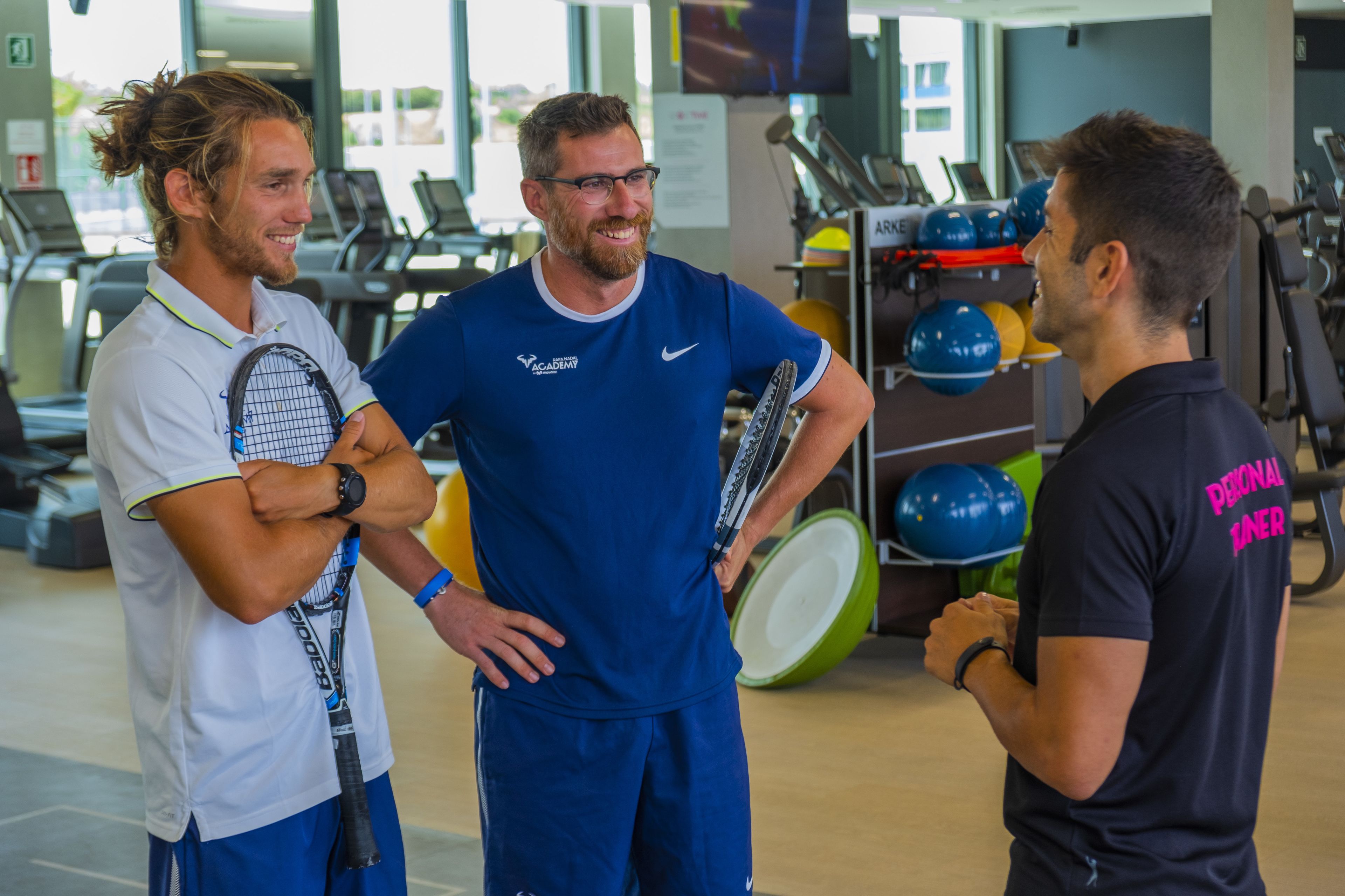 Trois personnes dans une salle de sport, devant des appareils d’exercice et un présentoir à boules de bowling.