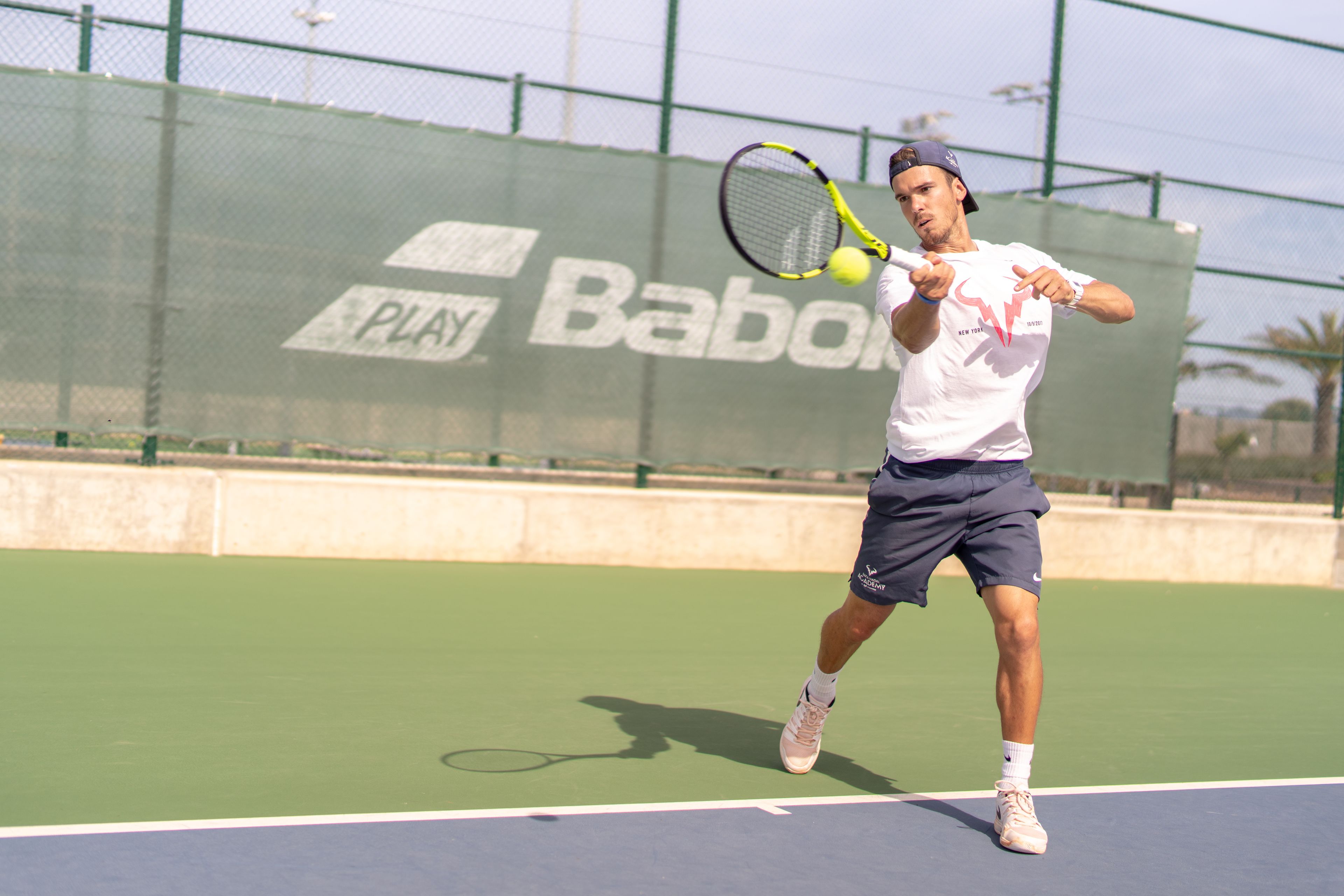 A tennis player on a court with a green surface, wearing a white shirt, dark shorts, and a cap.