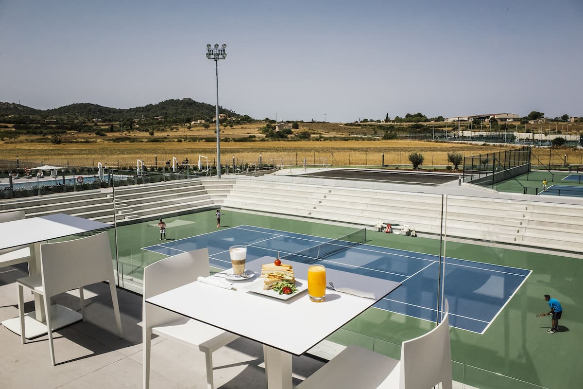 A tennis court with a table and chairs in the foreground, a person playing tennis, and a view of the surrounding landscape.