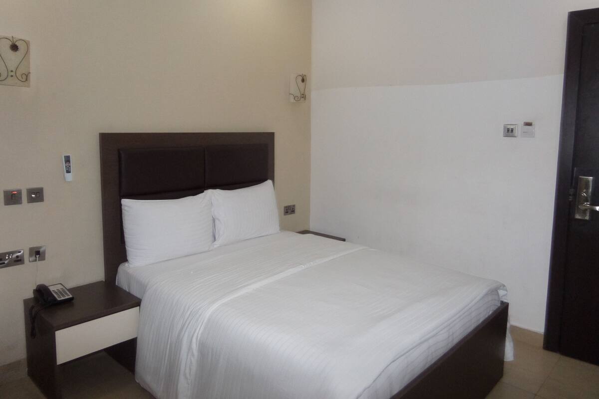 Una habitación de hotel con una cama individual, una mesita de noche, una lámpara fijada a la pared y una puerta.