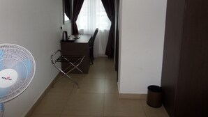 Una habitación pequeña y poco iluminada con un escritorio, una silla y un ventilador de pie.