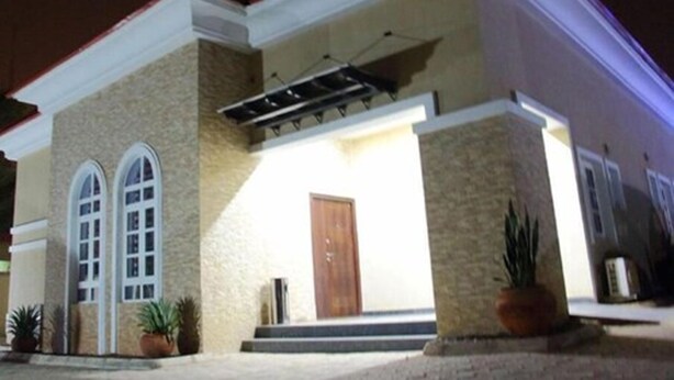 Un edificio de dos pisos con fachada color beige, una puerta de madera y un porche cubierto.
