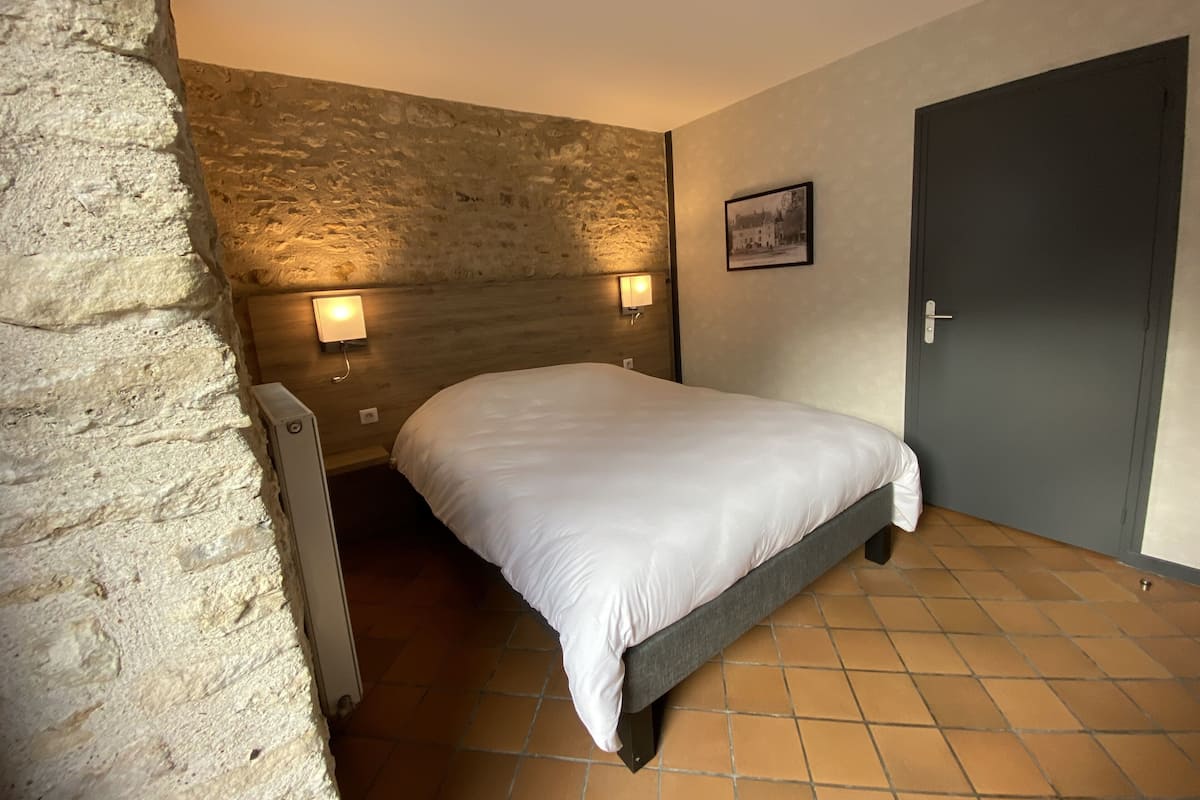 A bedroom with a stone wall, a bed with white bedding, two wall-mounted lights, and a door.