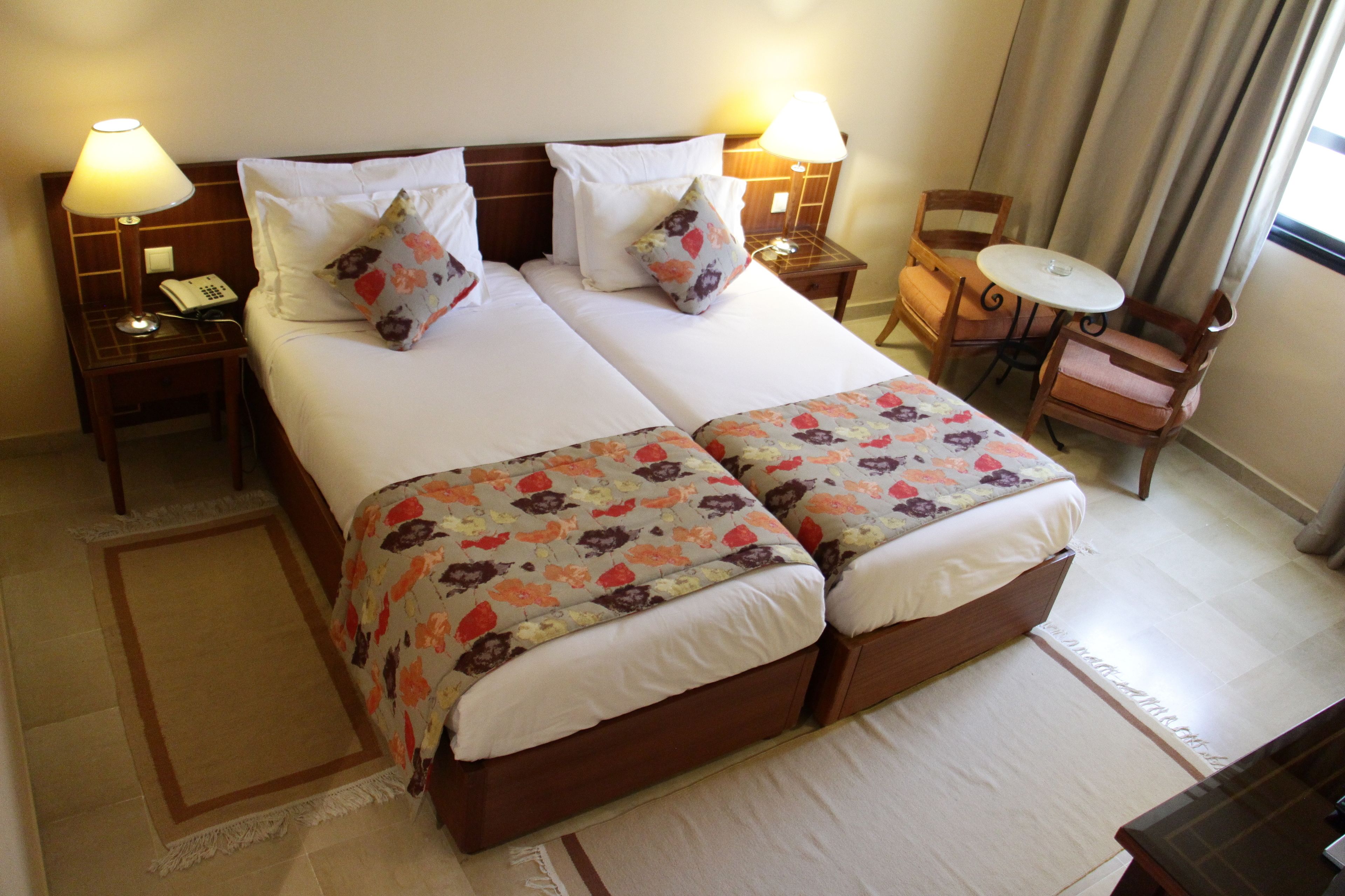 A double bed with floral bedding, two bedside tables with lamps, a small round table with chairs, and a view of the outdoors through a window.