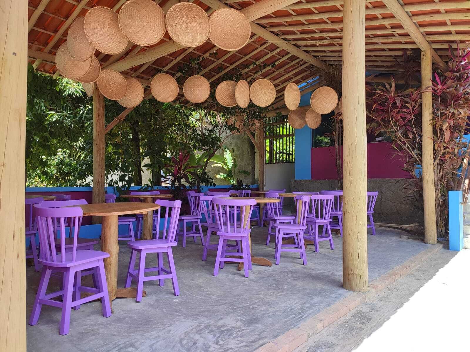 An outdoor seating area with wooden tables and purple chairs under a wooden pergola with woven baskets hanging from the ceiling.