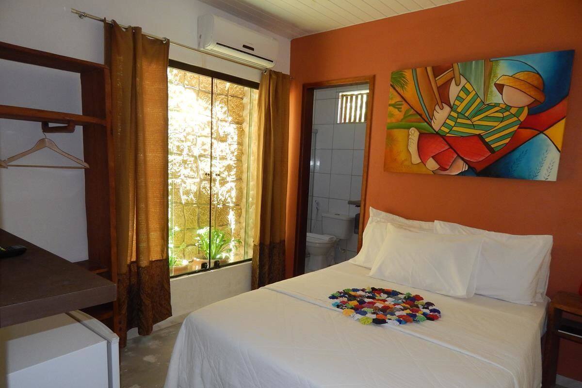 A hotel room with a bed, a window with curtains, a painting on the wall, and a small table with a chair.