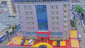 A multi-story building with a central glass facade, surrounded by a parking area with yellow and red markings. - Noubou International Hotel (Douala)