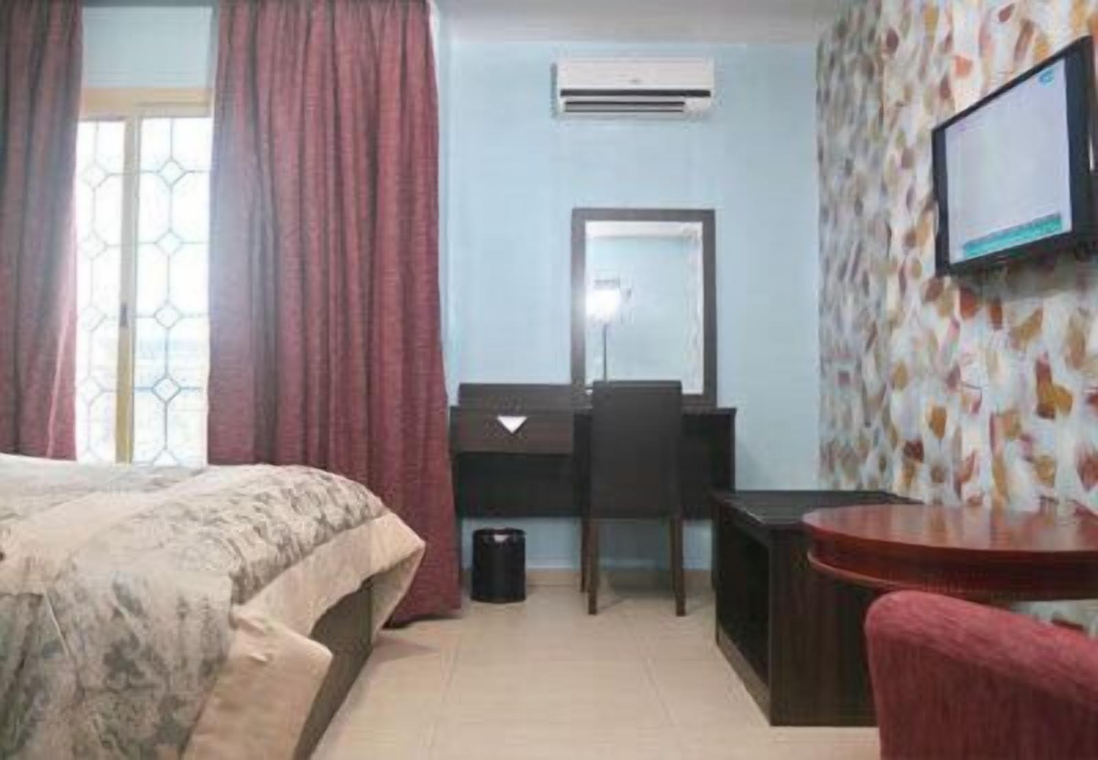 A hotel room with a bed, a desk, a chair, a TV, and an air conditioning unit.