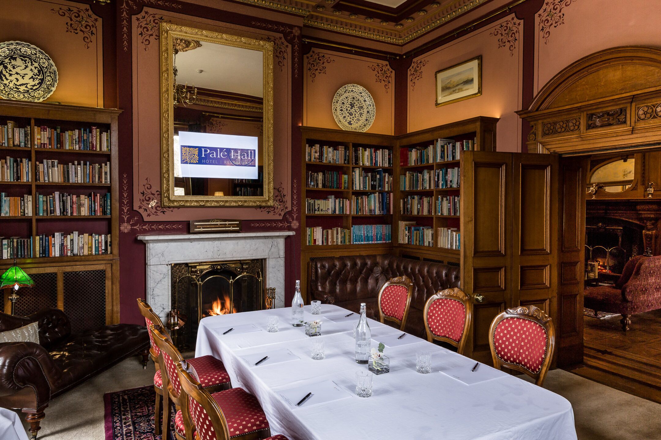 A library with bookshelves, a fireplace, a table set for dining, and a large mirror.