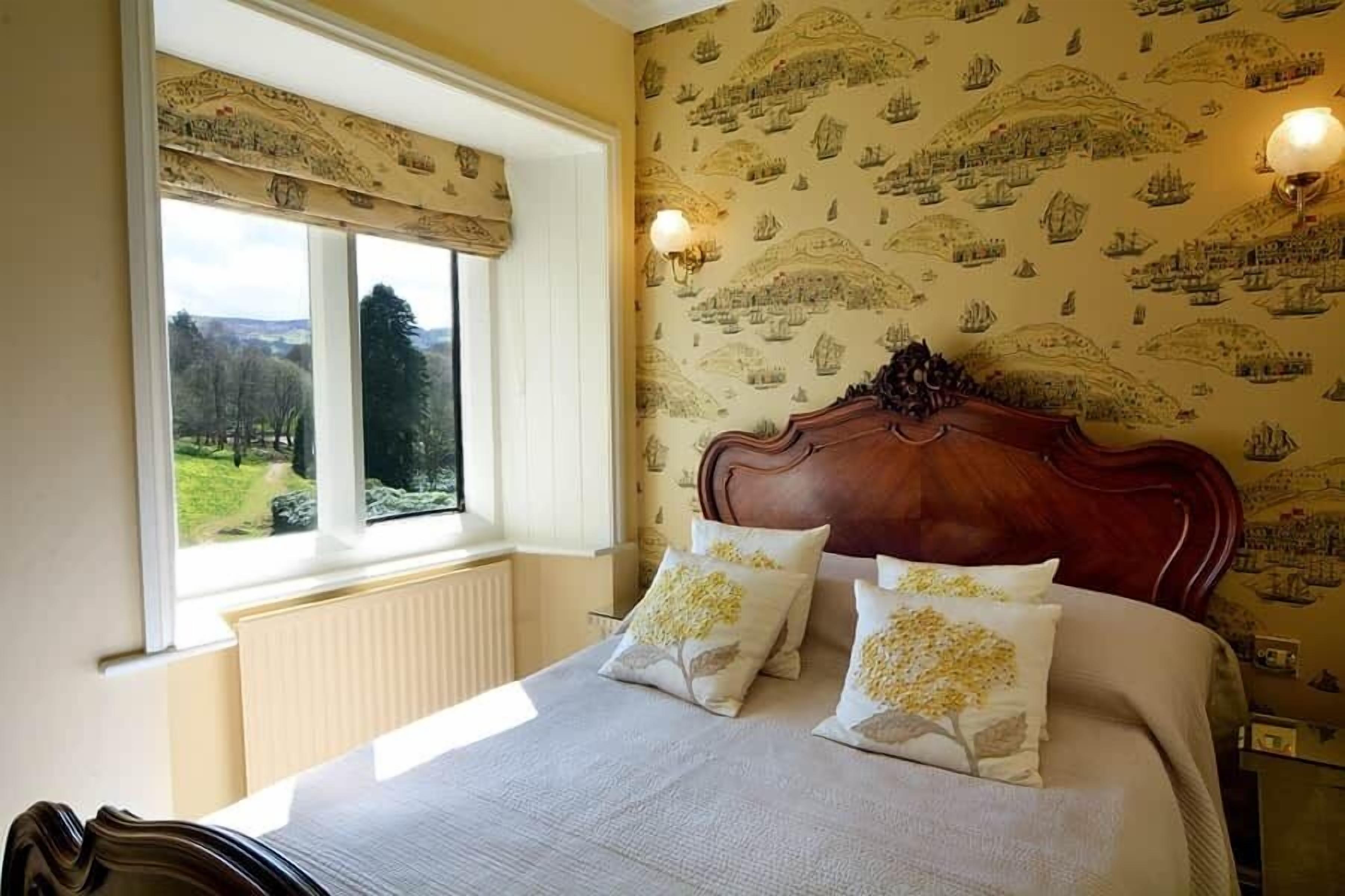 A bedroom with a large bed, a window with a view of greenery, and wallpaper with ship illustrations.