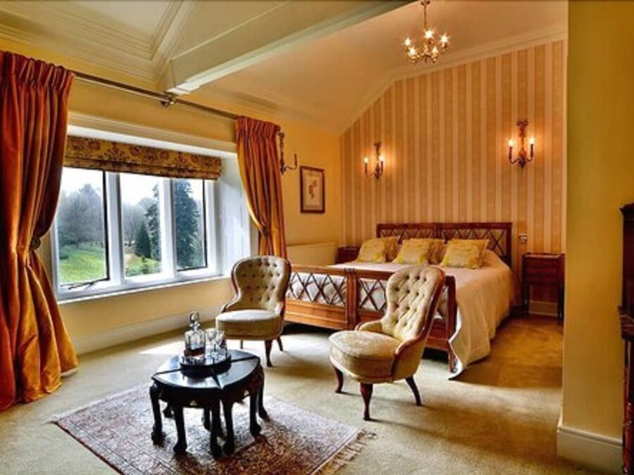 Een slaapkamer met een groot bed, twee fauteuils, een bijzettafeltje en uitzicht op het landschap door een raam.