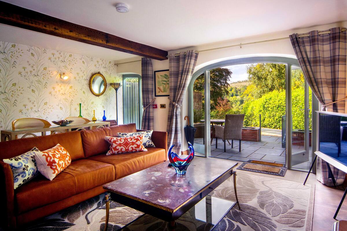 Un salon chaleureux avec un canapé en cuir marron, une table basse en verre et une vue sur un jardin, visible à travers une porte vitrée.