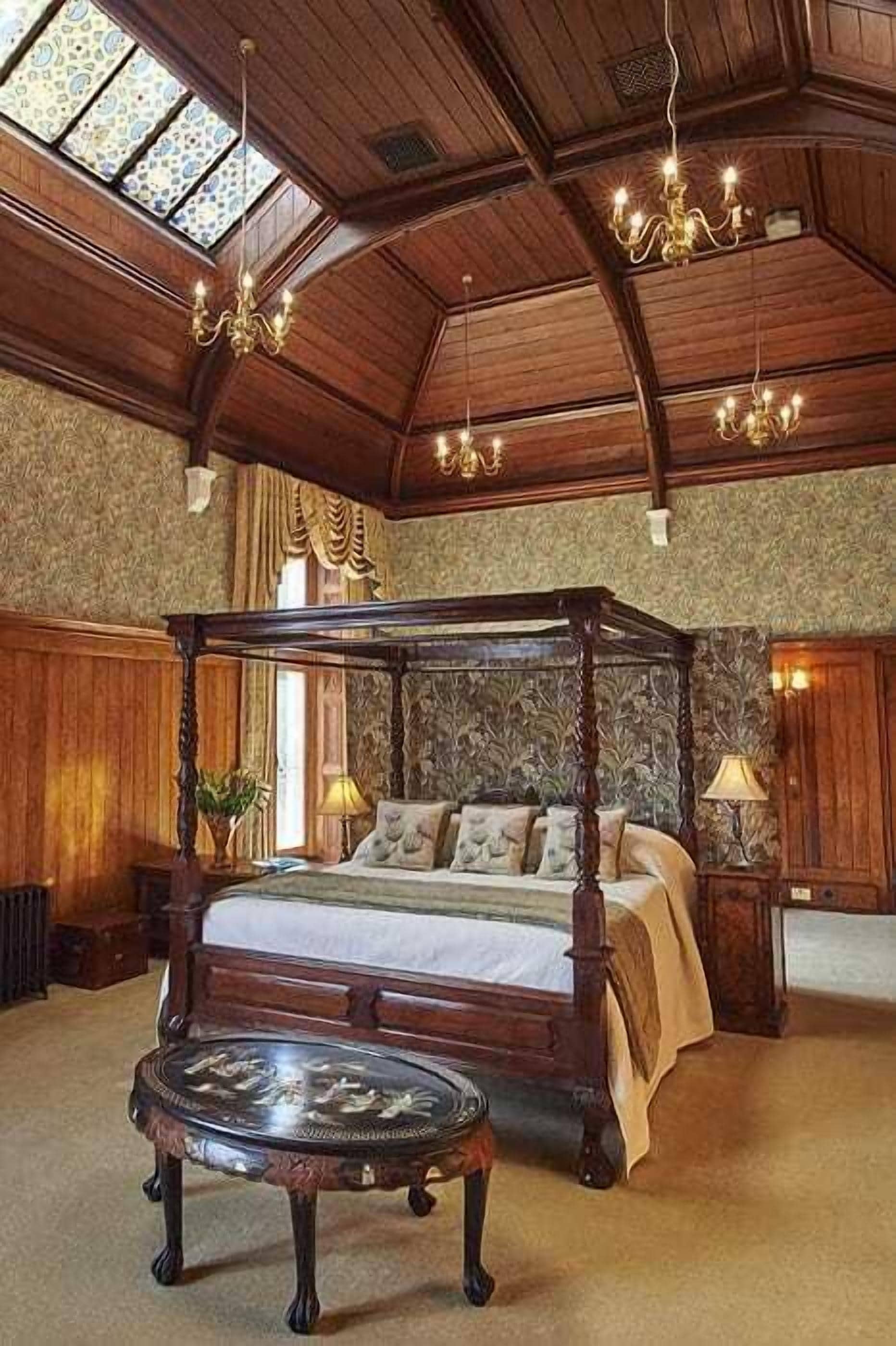 A four-poster bed with a canopy, a round table, and a chandelier in a room with wooden paneling and a high ceiling.