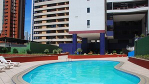 Una piscina con borde de baldosas, rodeada por un muro bajo y plantas. Al fondo, edificios de varios pisos con balcones.