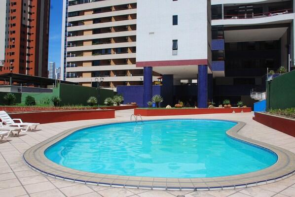 A swimming pool with a tiled deck, surrounded by a low wall and plants. In the background, there are multi-story buildings with balconies.