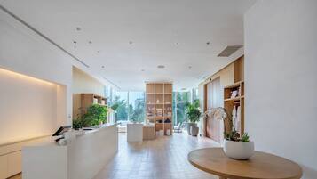 A modern hotel lobby with a reception desk, wooden shelving, potted plants, and a view of the city.