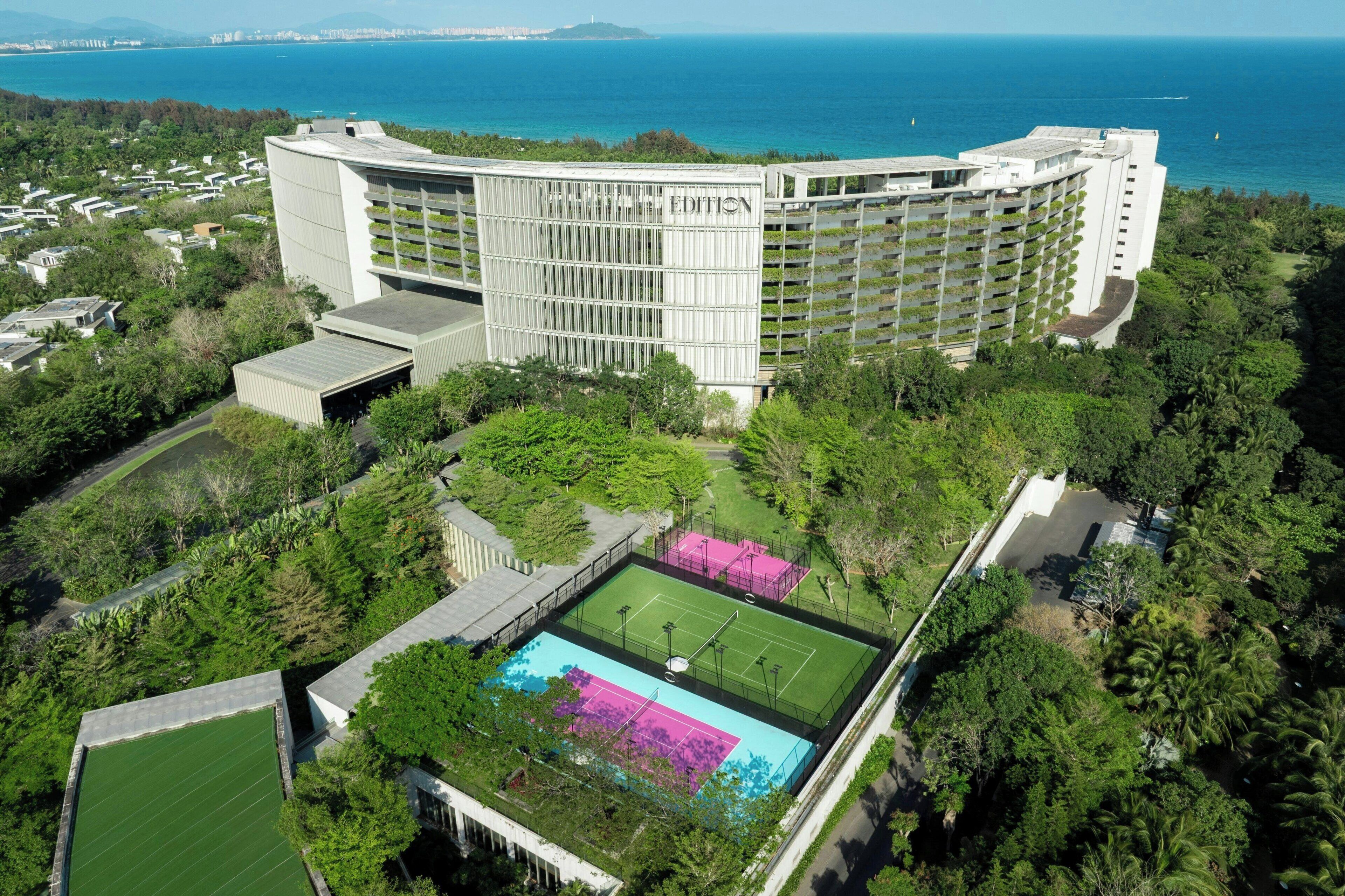 A large hotel with multiple floors, surrounded by lush greenery and a tennis court.
