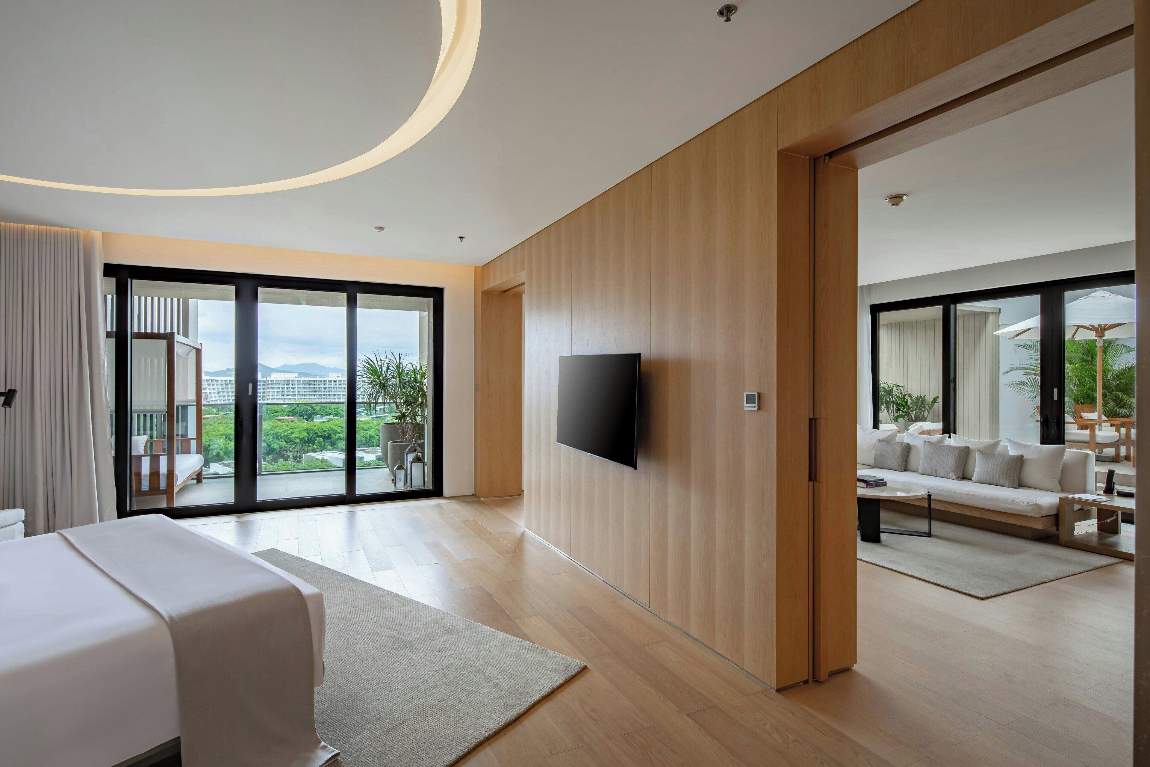 Una habitación de hotel moderna con una cama grande, un televisor de pantalla plana, paneles de madera, una puerta de vidrio corredera a un balcón y una zona de estar con sofá y mesa de centro.