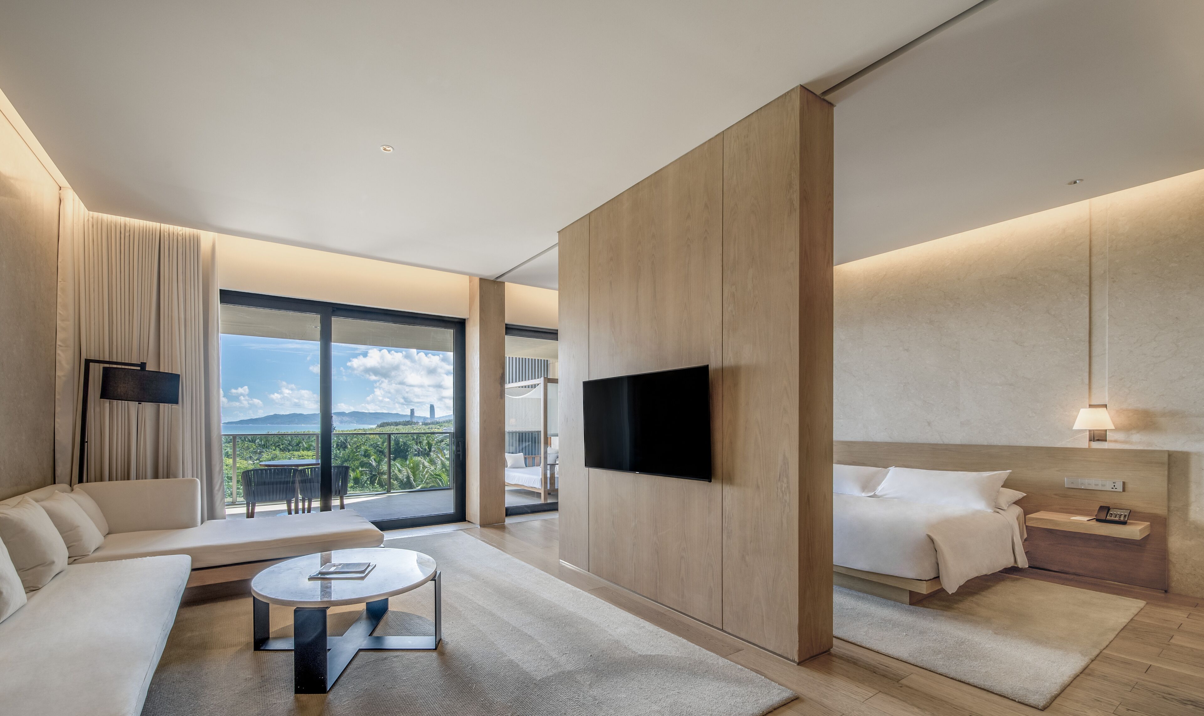A modern hotel room with a sofa, a round coffee table, a bed, a TV mounted on the wall, and a view of the outdoors.