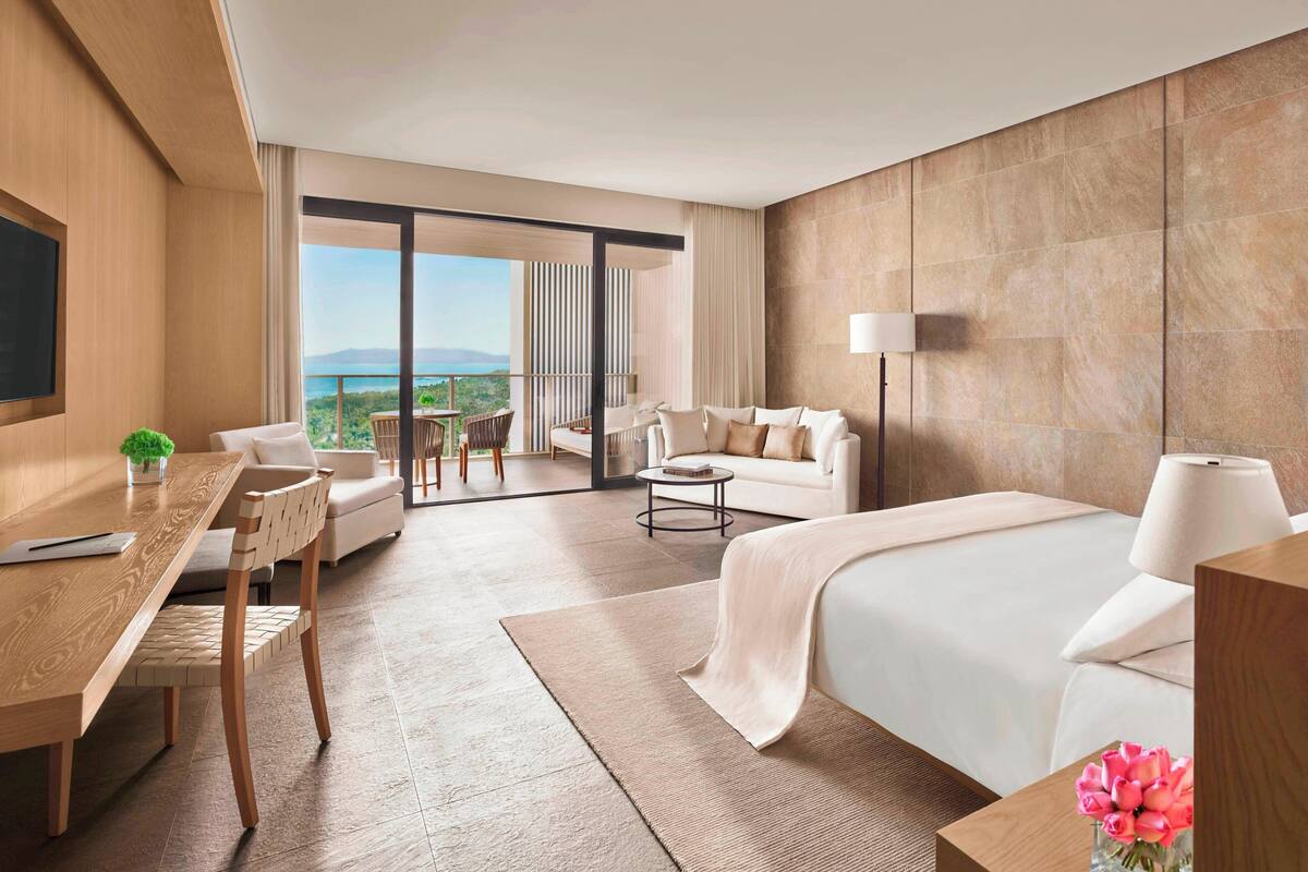 A modern hotel room with a large bed, a desk, a sofa, and a balcony with a view.
