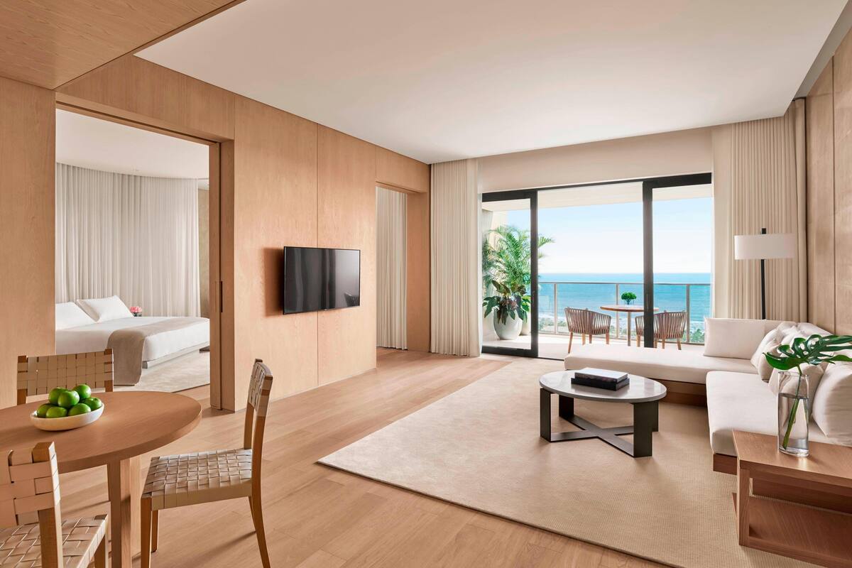 A modern hotel room with a large balcony, a flat-screen TV, a dining area with a table and chairs, and a sofa with a view of the sea.