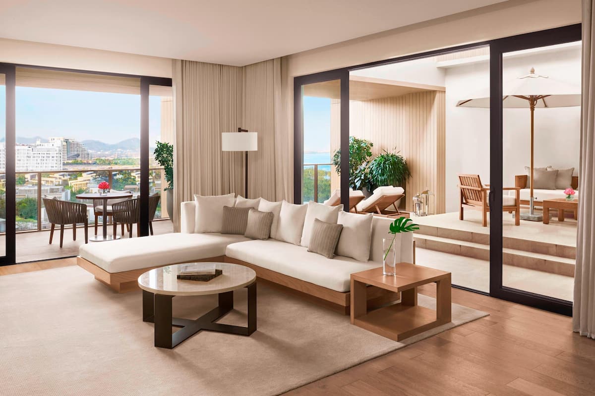 A modern living room with a large sectional sofa, a coffee table, and a dining area with chairs and a table. Large windows offer a view of the city and mountains.