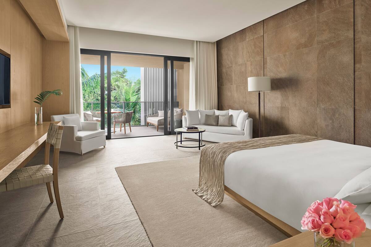 A modern hotel room with a large bed, a desk, a sofa, and a balcony with a view of greenery.