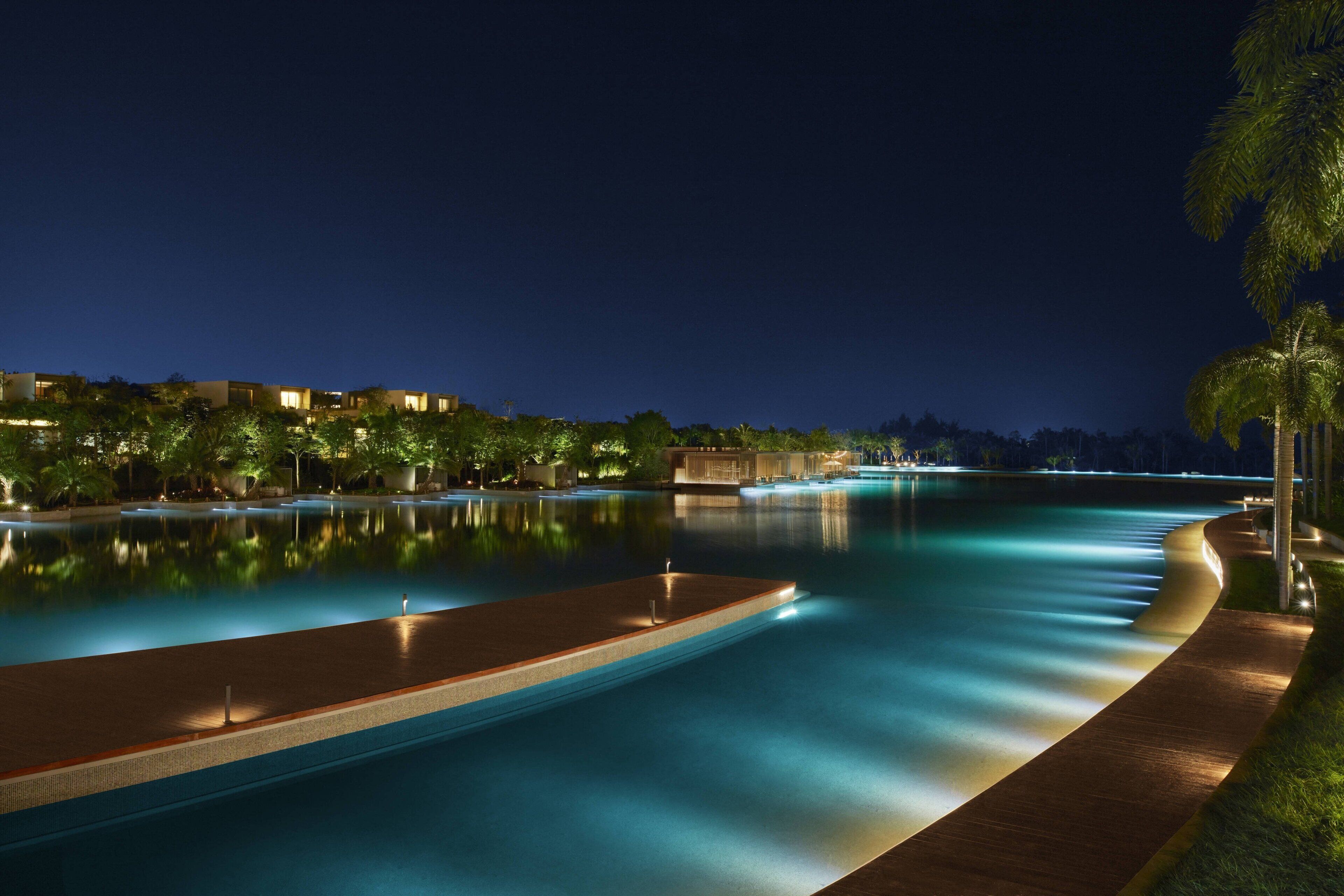 A nighttime view of a swimming pool with a wooden deck and palm trees.