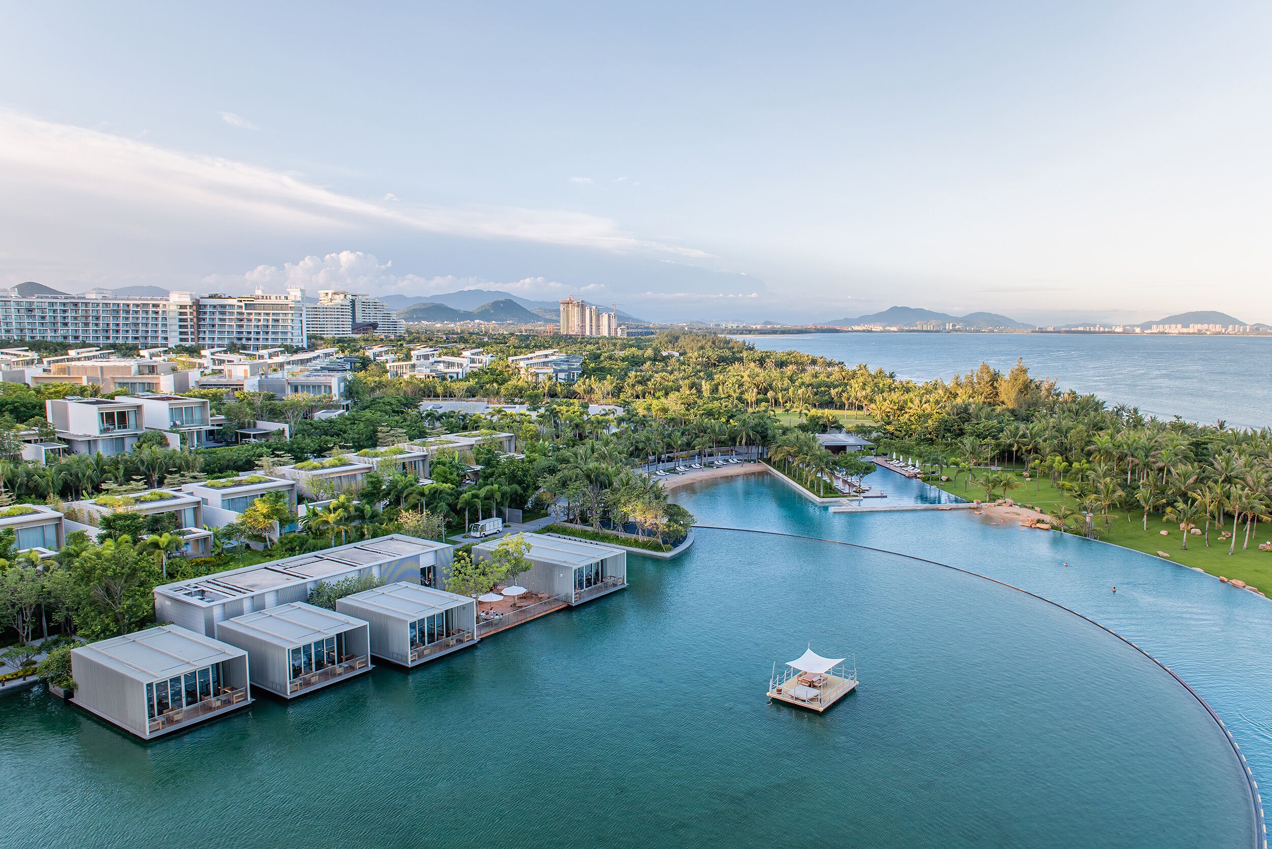 Un hôtel moderne situé au bord de l’eau, doté de villas flottantes, d’un port de plaisance et offrant une vue sur la mer et les montagnes lointaines.