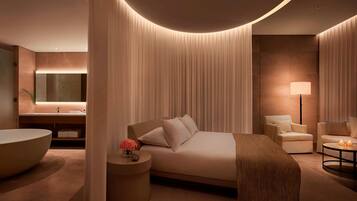 A modern hotel room with a large bed, a sitting area with a chair, a round coffee table, a large bathtub, and a vanity with a mirror and sink.