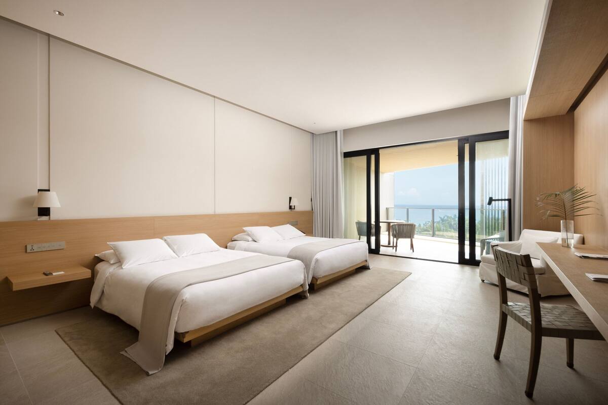 A modern hotel room with two beds, a desk, and a balcony with a view.