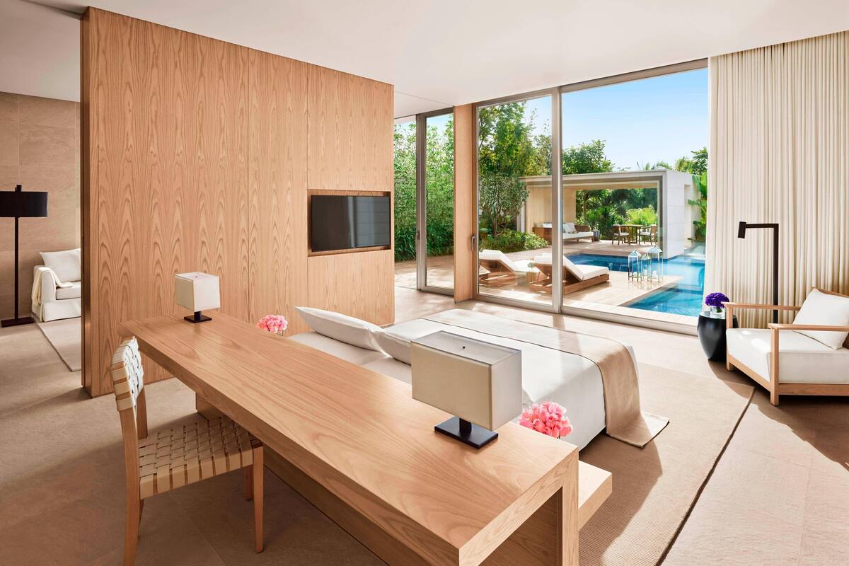 A modern hotel room with a wooden desk, a flat-screen TV, a bed with a white headboard, and a large window overlooking a pool and garden.