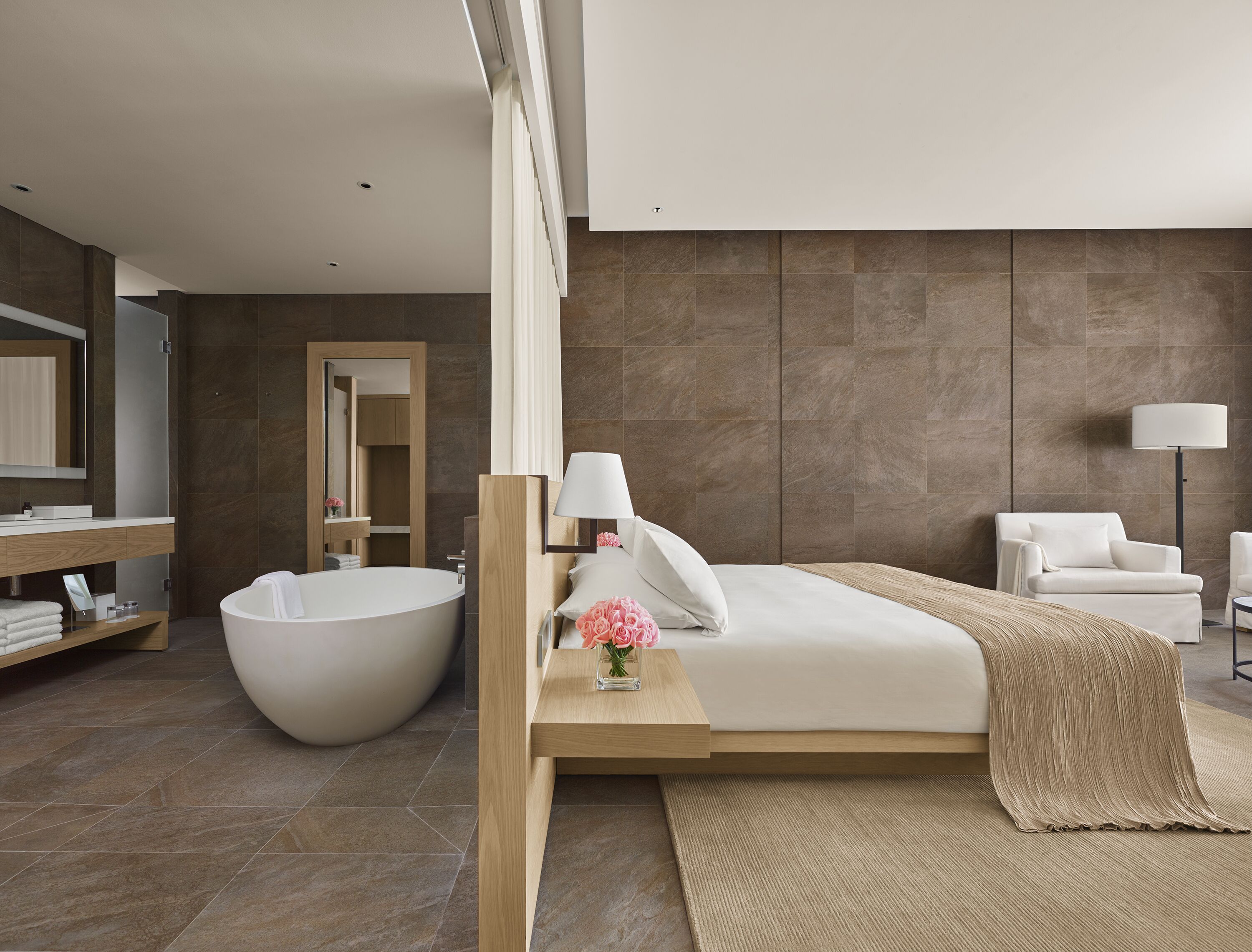 Ein modernes Hotelzimmer mit einer großzügigen Badewanne, einem hölzernen Bettgestell, einem Nachttisch mit Lampe sowie einem Sessel.