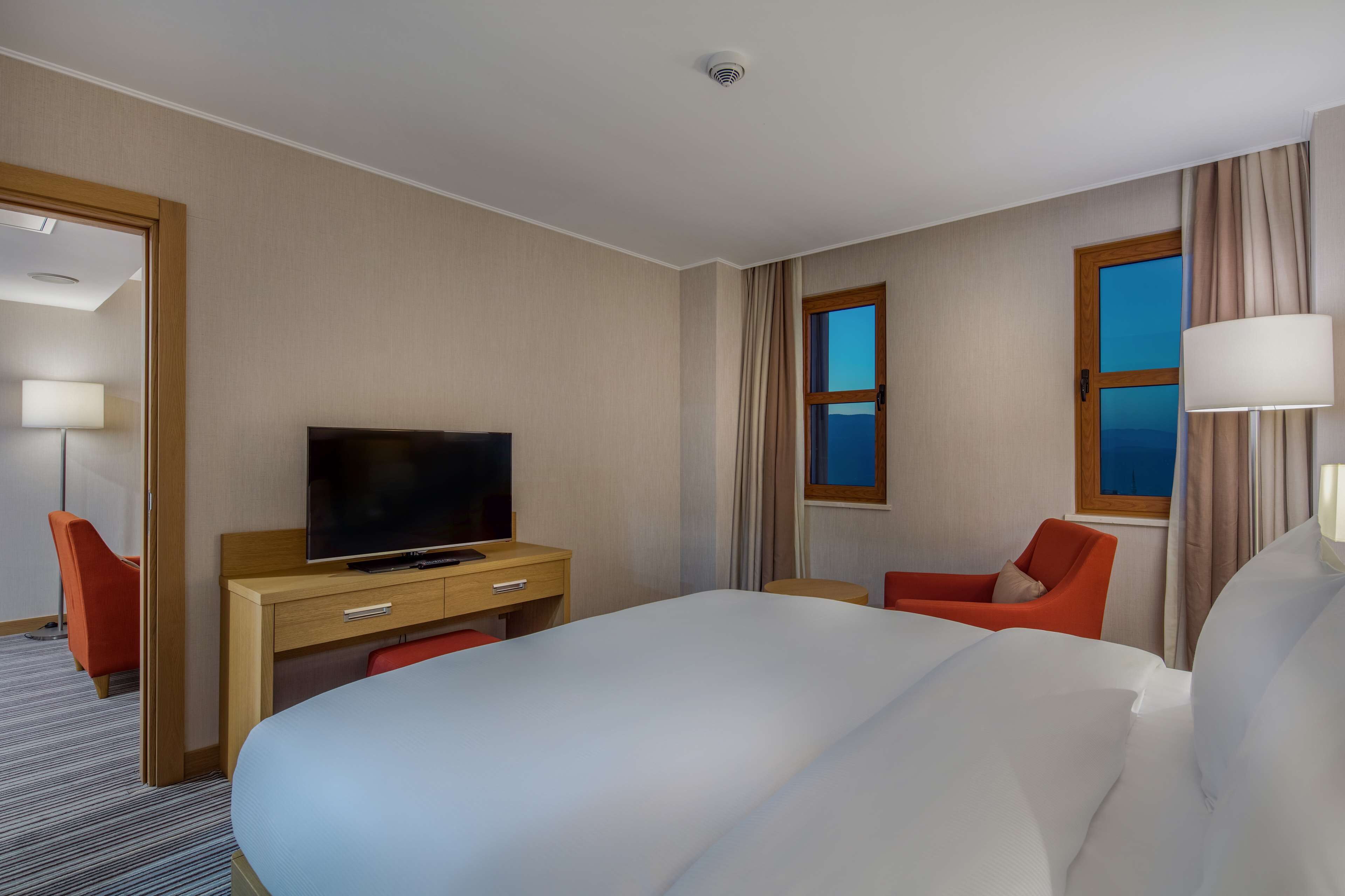 A hotel room with a large bed, a desk with a television, two chairs, and a window with curtains.