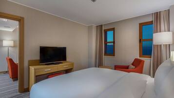 A hotel room with a large bed, a desk with a television, two chairs, and a window with curtains.