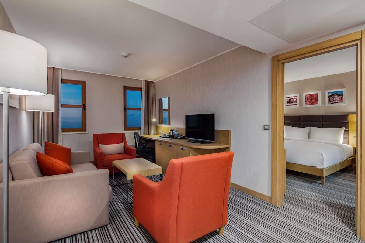 A hotel room with a sofa, armchair, desk, TV, and a bed.