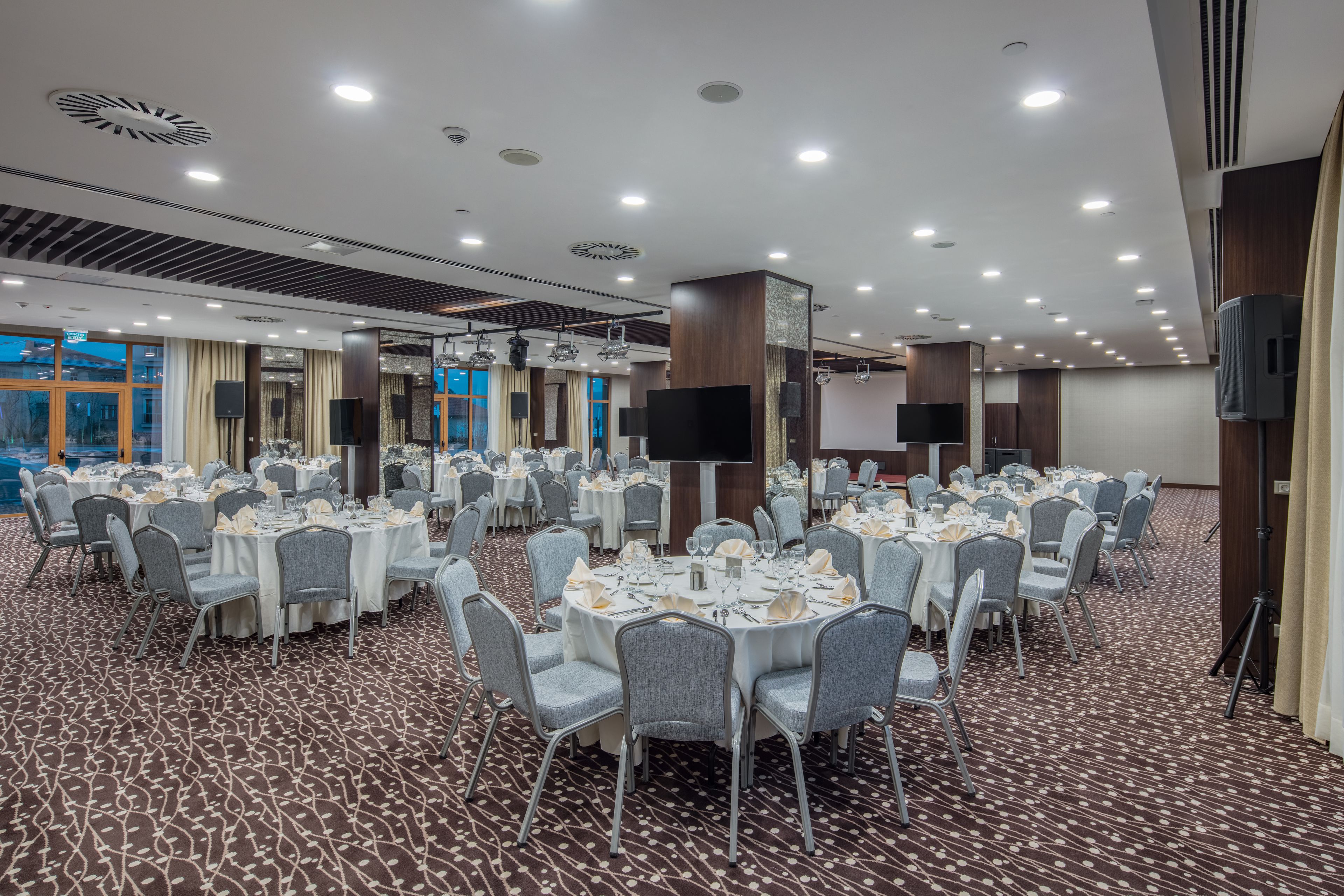 A large conference room with round tables set for dining, equipped with chairs, tables, and a large screen.