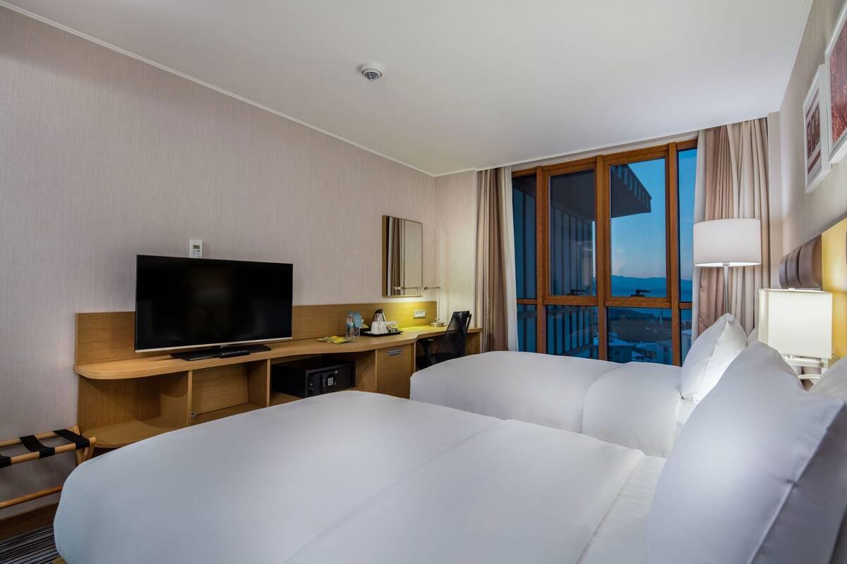 A hotel room with a bed, a TV, a desk, a lamp, and a window with a view.