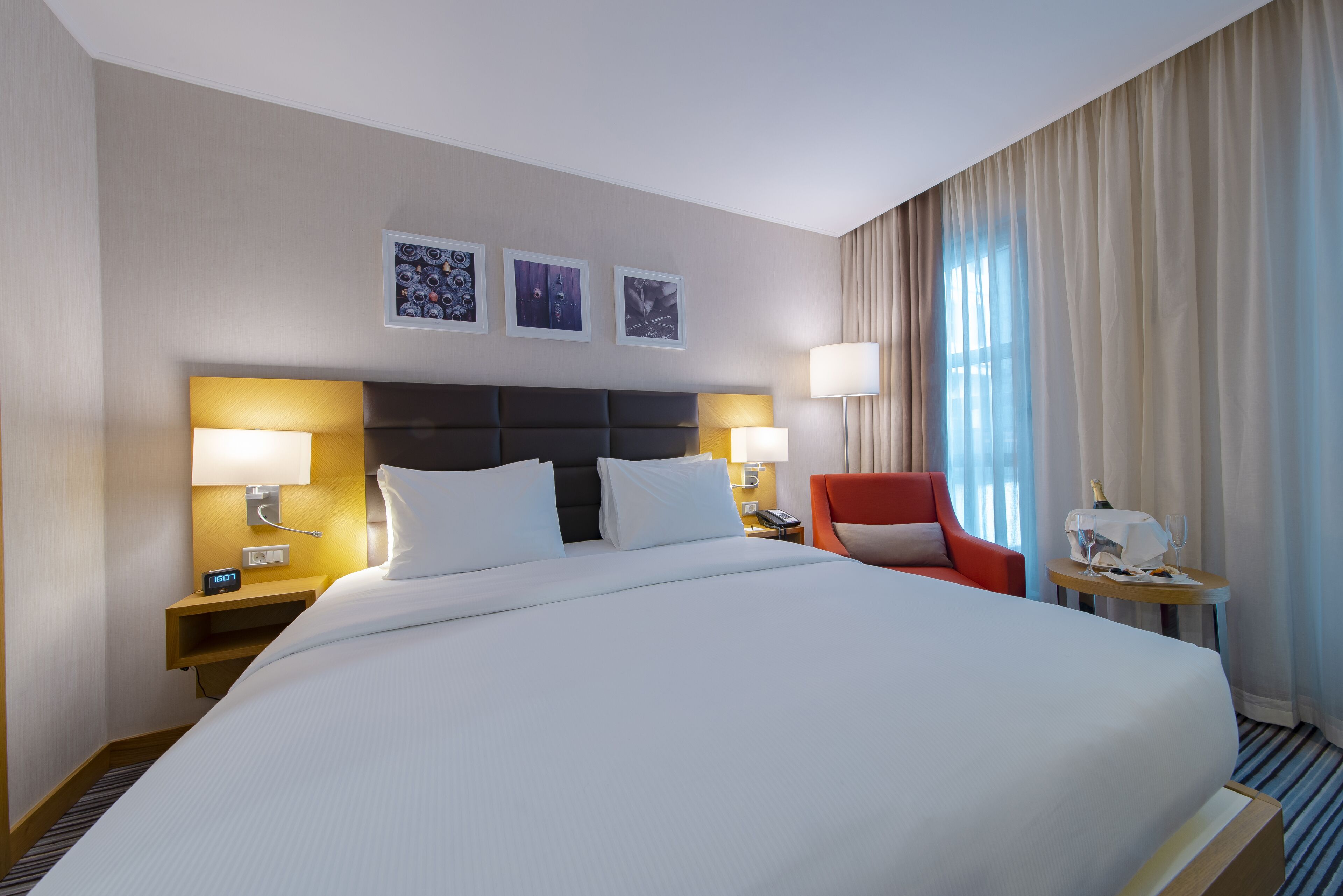 Een hotelkamer met een groot bed, twee nachtkastjes met lampen, een rode fauteuil en een bijzettafeltje met een fles en glazen.