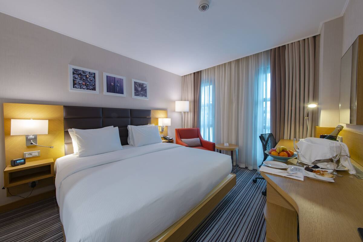 A hotel room with a large bed, two bedside tables with lamps, a red chair, a small table with fruit and a bottle, and a large window with curtains.
