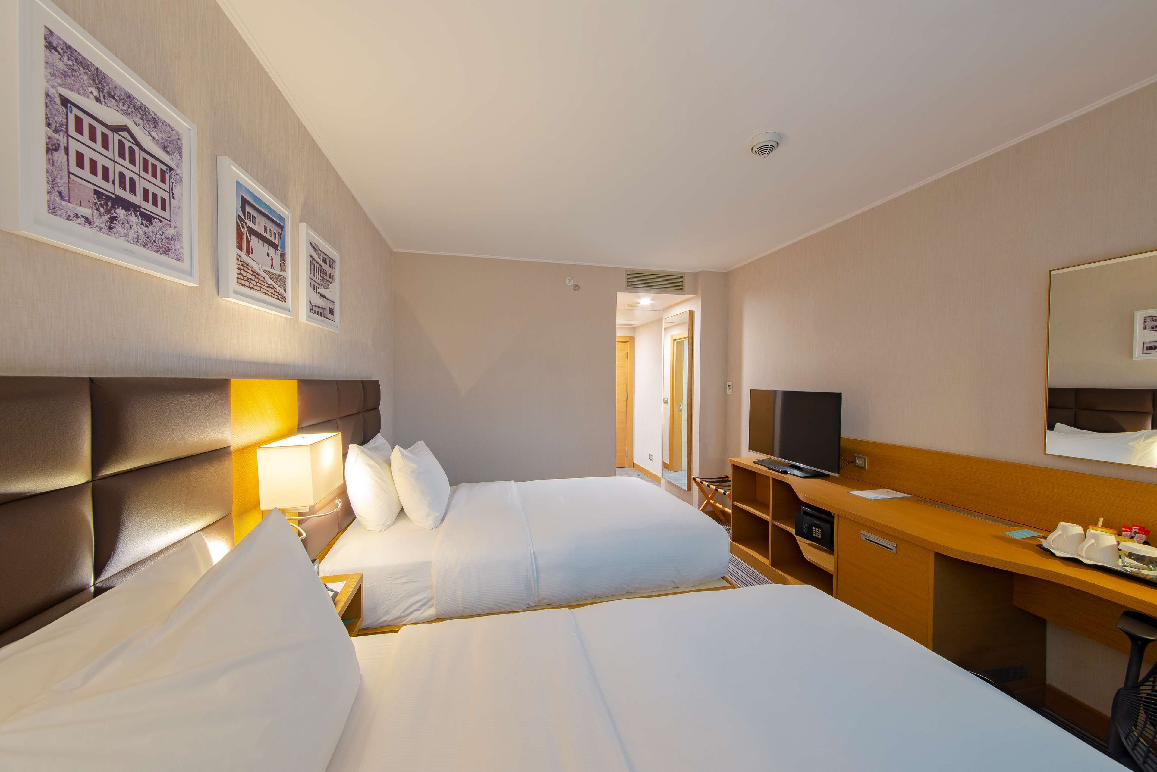 A hotel room with two beds, a desk, a television, and framed pictures on the wall.