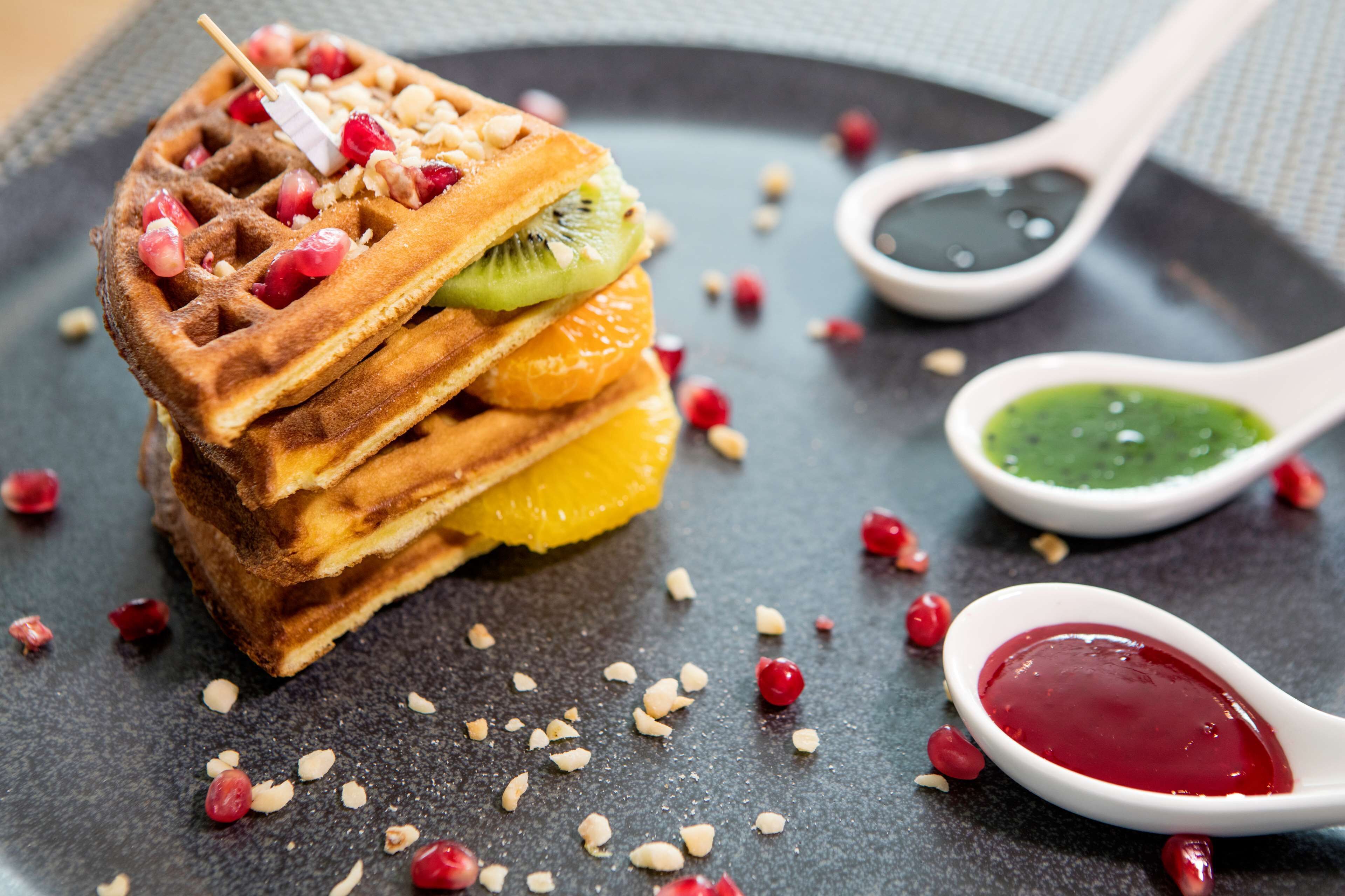 A stack of waffles with fruit and nuts, accompanied by two small bowls of green and red sauces on a dark plate.