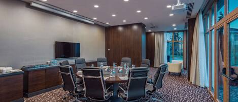 A conference room with a round table, chairs, a TV, and a window with curtains.