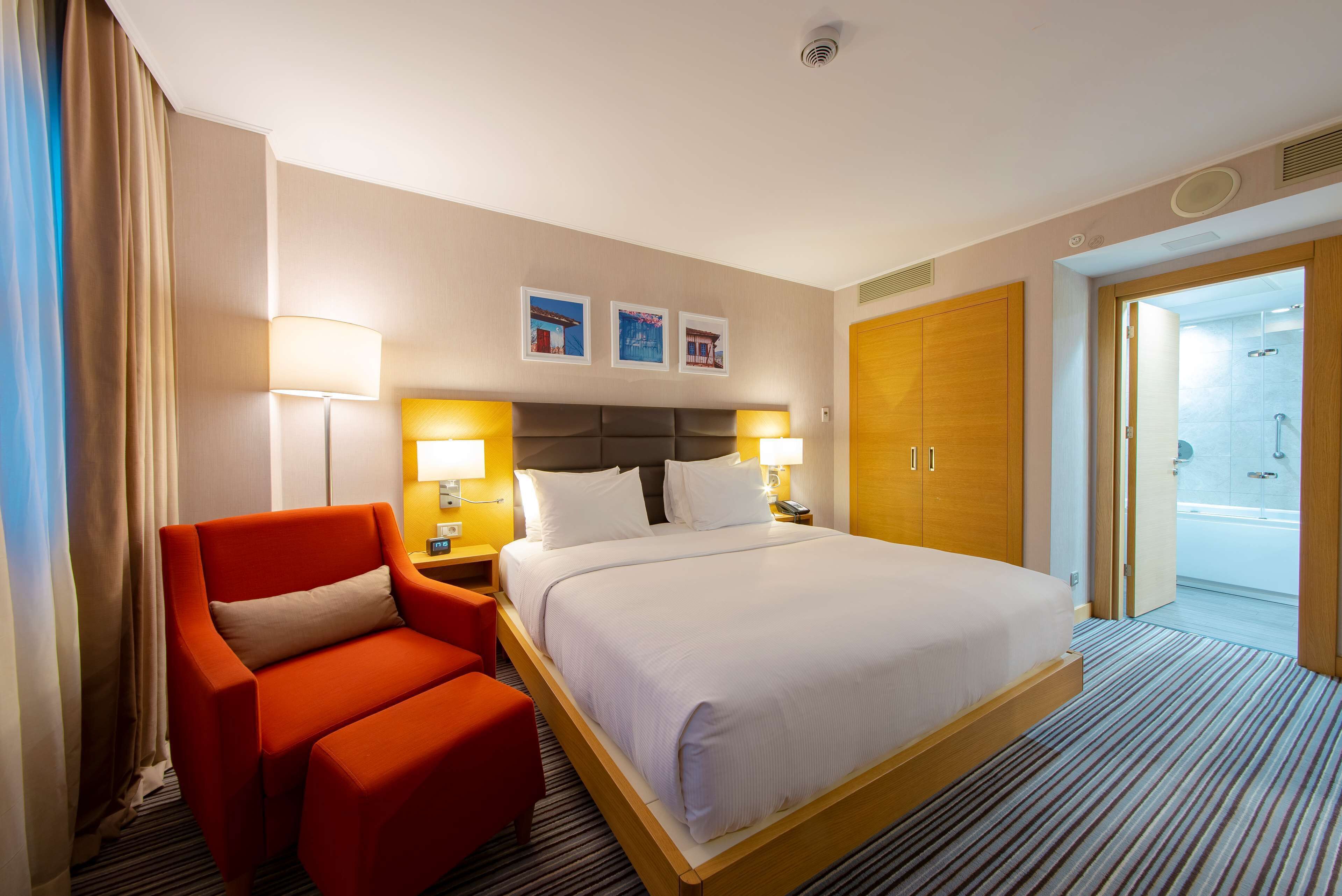 A hotel room with a large bed, a red armchair, a nightstand with a lamp, and a wooden wardrobe.