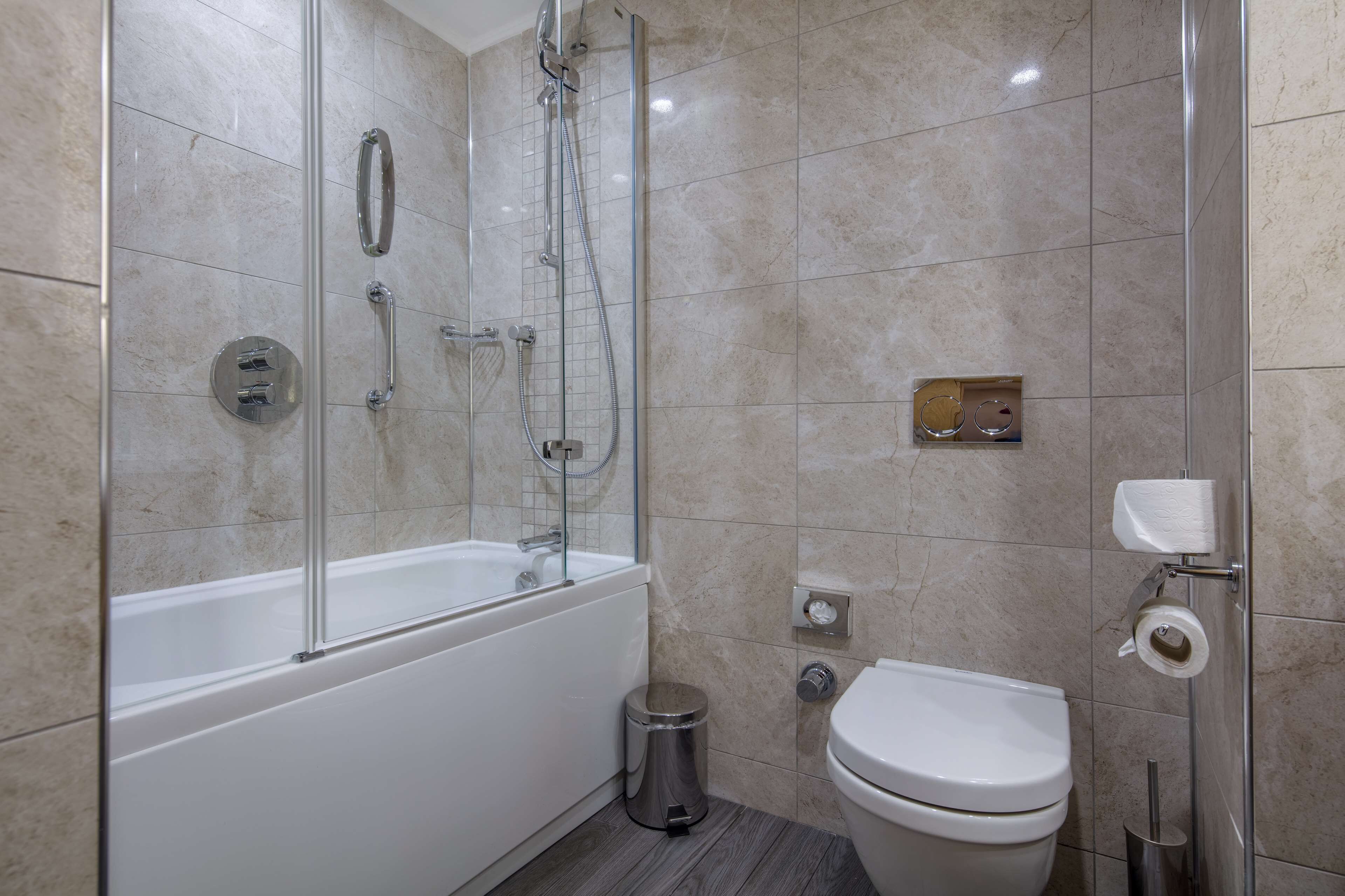 A modern bathroom with a glass-enclosed shower, a white bathtub, a toilet, and a trash can.