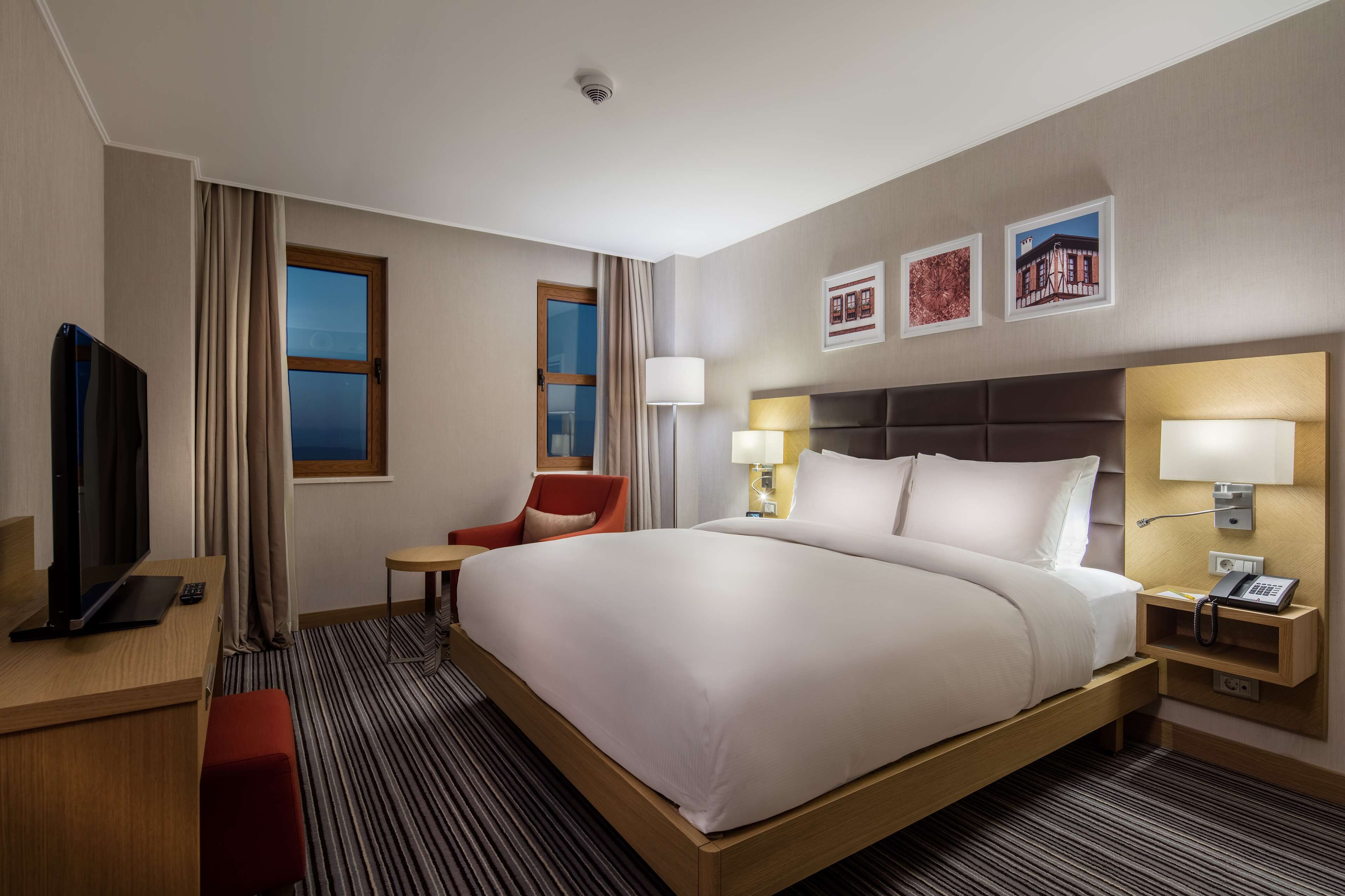 A hotel room with a large bed, a desk with a TV, a chair, and a bedside table with a lamp.