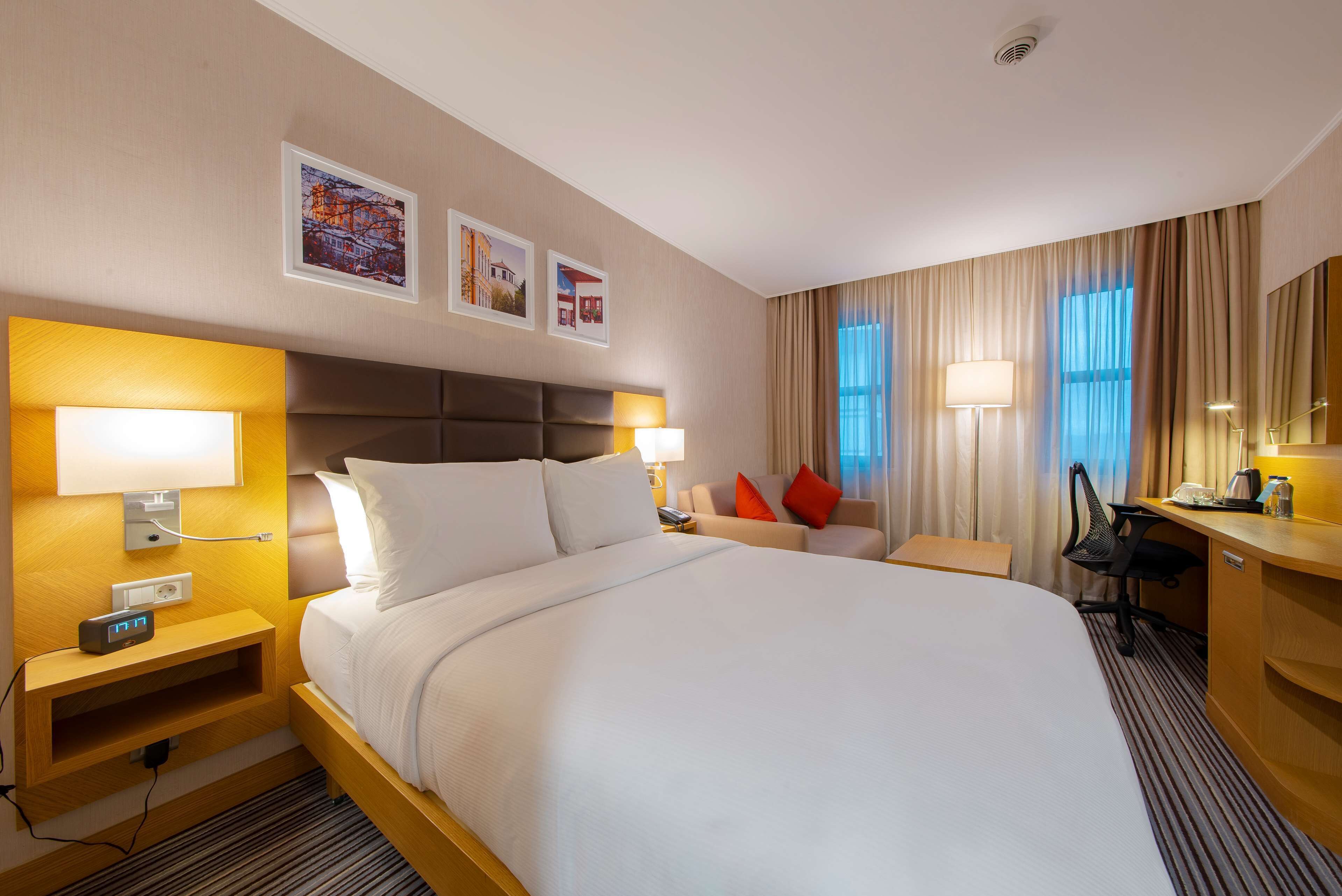 Habitación de hotel con una cama grande, mesitas de noche con lámparas, un escritorio y una silla. Se observan cuadros enmarcados en la pared y una ventana con cortinas.